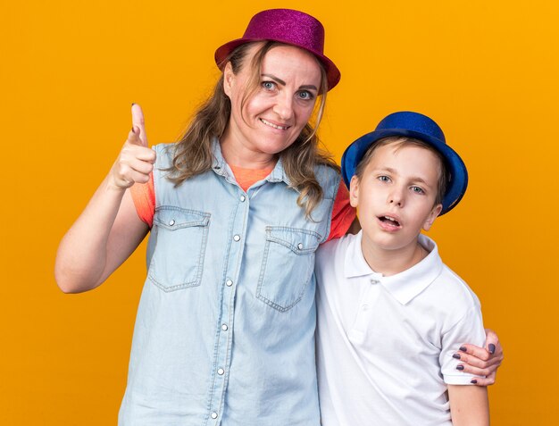 verärgerter junger slawischer Junge mit blauem Partyhut, der mit seiner Mutter steht, die einen lila Partyhut trägt und auf die Seite zeigt, die auf oranger Wand mit Kopienraum isoliert ist?