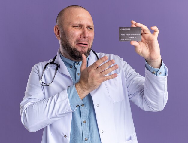 Verärgerter männlicher Arzt mittleren Alters, der ein medizinisches Gewand und ein Stethoskop trägt und die Kreditkarte hält und darauf zeigt
