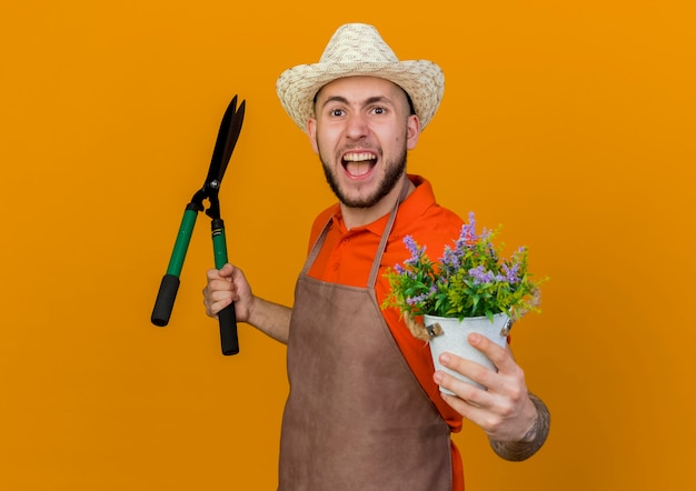 Verärgerter männlicher Gärtner, der Gartenhut trägt, hält Haarschneidemaschinen und Blumen im Blumentopf