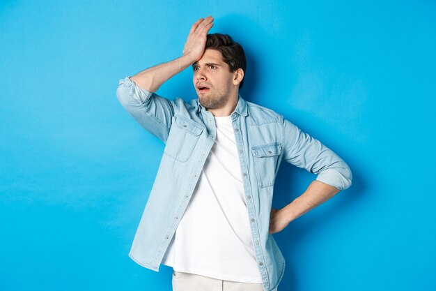 Verärgerter mann, der facepalm macht und besorgt wegschaut, etwas wichtiges vergessen, beunruhigt auf die stirn schlagen, vor blauem hintergrund stehen