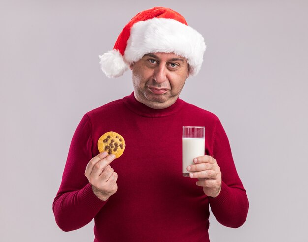 Verärgerter Mann mittleren Alters mit Weihnachtsmütze, der ein Glas Milch und Kekse mit traurigem Ausdruck über der weißen Wand hält