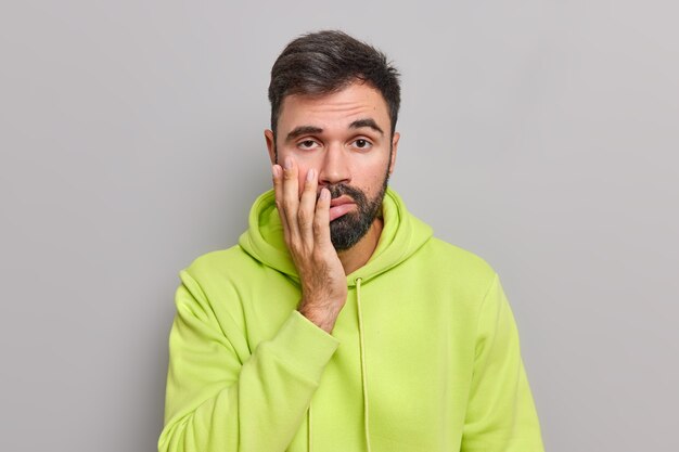 Verärgerter müder Mann hält die Hand auf dem Gesicht hat desinteressierten gelangweilten Gesichtsausdruck, der von langweiligen Gesprächen satt ist, trägt ein grünes Sweatshirt