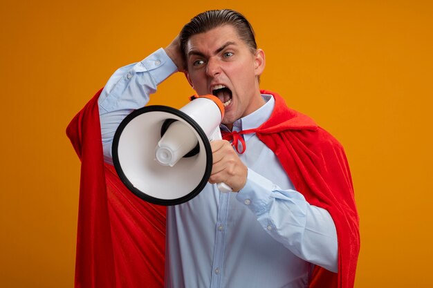 Verärgerter Superheld-Geschäftsmann im roten Umhang, der zum Megaphon mit aggressivem Ausdruck schreit, der über orange Wand steht
