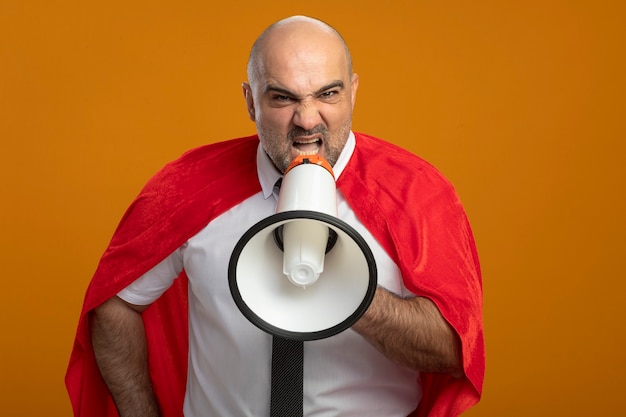 Verärgerter Superheld-Geschäftsmann im roten Umhang, der zum Megaphon mit aggressivem Ausdruck schreit, der über orange Wand steht