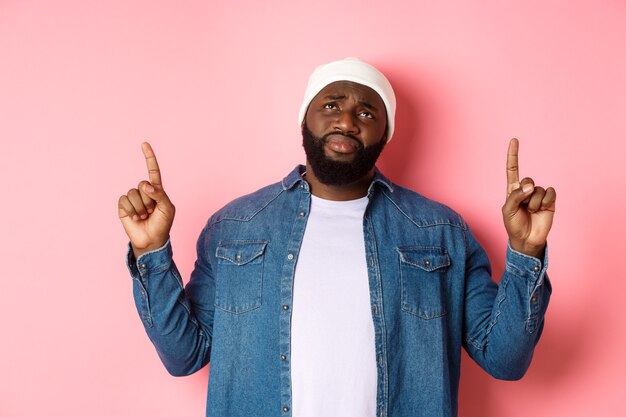 Verärgerter und düsterer afroamerikanischer Mann in Mütze, der mit den Fingern nach oben zeigt, seufzt und mit Traurigkeit nach oben schaut und auf rosafarbenem Hintergrund steht