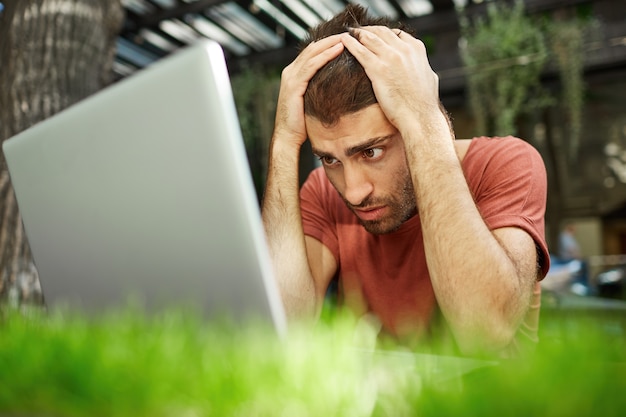 Verärgerter und enttäuschter gutaussehender Kerl, der traurig auf Laptop-Bildschirm schaut, während er draußen sitzt und während covid-19 an der Fernbedienung arbeitet