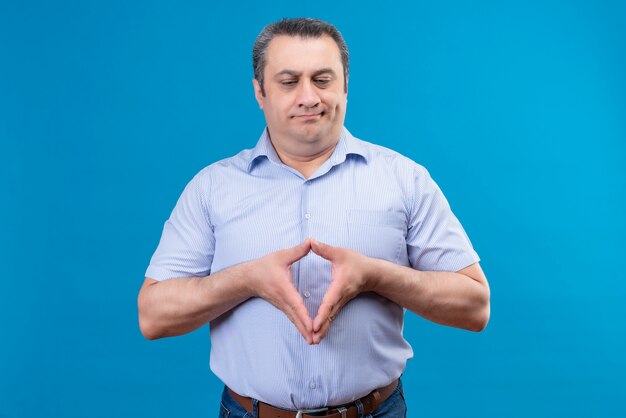 Verärgerter und verwirrter Mann mittleren Alters in blau gestreiftem Hemd, der an einem blauen Raum denkt und Hände zusammenhält