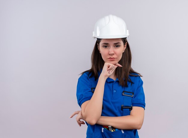 Verärgertes junges Baumeistermädchen mit weißem Schutzhelm und blauer Uniform legt Hand auf Kinn und betrachtet Kamera auf lokalisiertem weißem Hintergrund mit Kopienraum