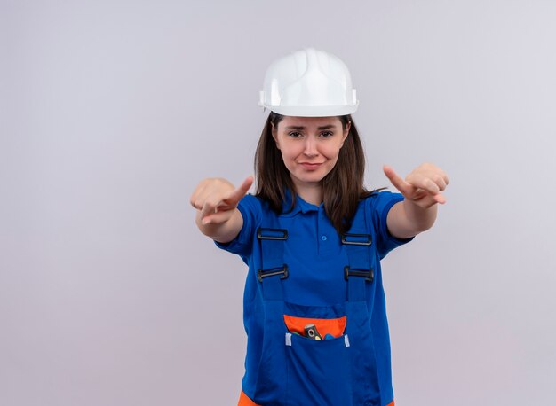 Verärgertes junges Baumeistermädchen mit weißem Schutzhelm und blauer Uniform zeigt mit beiden Händen auf lokalem weißem Hintergrund mit Kopienraum nach vorne