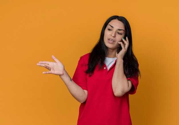 Verärgertes junges brünettes kaukasisches Mädchen, das rotes Hemd trägt, spricht am Telefon und zeigt auf Seite, die auf orange Wand lokalisiert wird