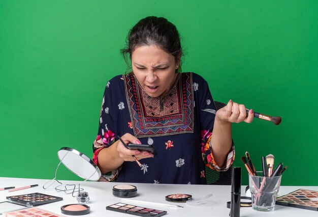 Verärgertes junges brünettes Mädchen, das am Tisch mit Make-up-Tools sitzt und Make-up-Pinsel hält und auf das Telefon isoliert auf grüner Wand mit Kopierraum schaut