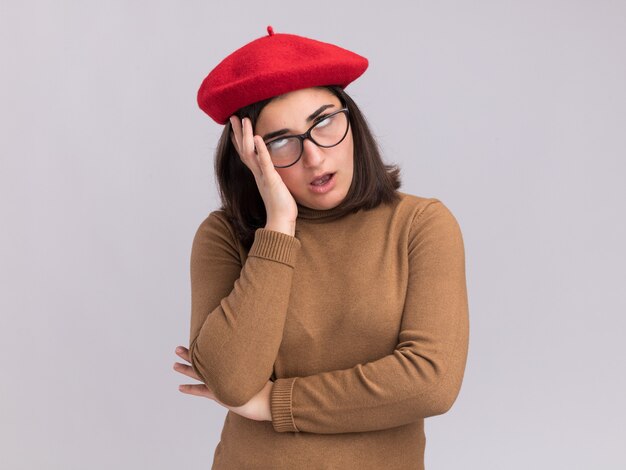 Verärgertes junges hübsches kaukasisches Mädchen mit Baskenmütze und in optischen Gläsern rollende Augen und Hand auf Gesicht