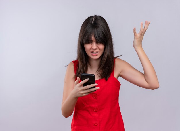 Verärgertes junges kaukasisches Mädchen, das Telefon mit erhabener Hand hält, die Telefon auf lokalisiertem weißem Hintergrund mit Kopienraum betrachtet