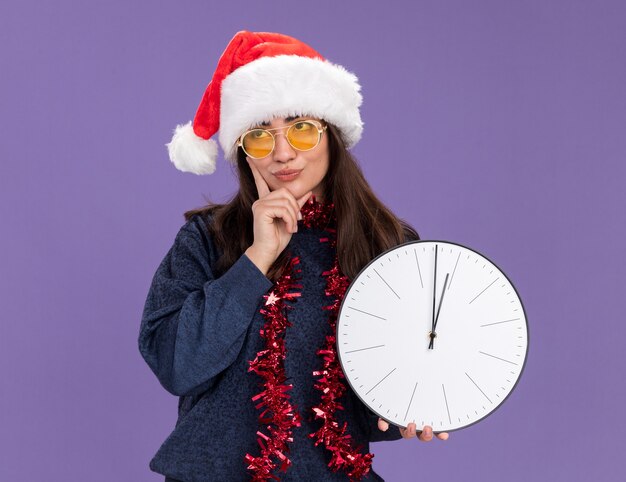 Verärgertes junges kaukasisches Mädchen in Sonnenbrille mit Weihnachtsmütze und Girlande um den Hals hält die Uhr und legt die Hand auf das Kinn rollende Augen einzeln auf lila Wand mit Kopierraum