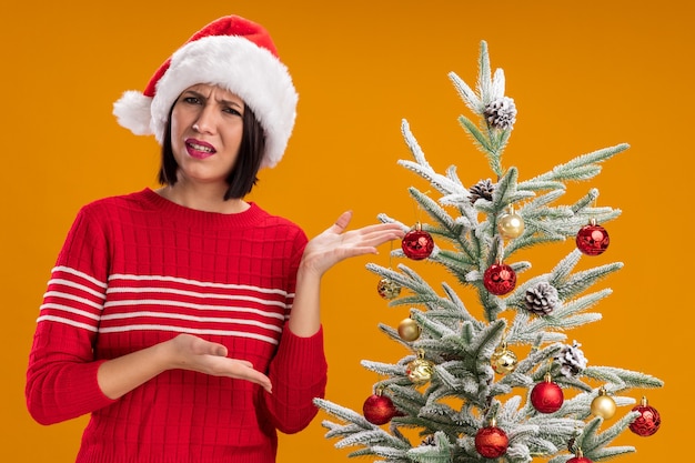 Verärgertes junges Mädchen mit Weihnachtsmütze, das in der Nähe des geschmückten Weihnachtsbaums steht und auf die Kamera blickt, die mit den Händen auf den Baum zeigt, der auf orangefarbenem Hintergrund isoliert ist