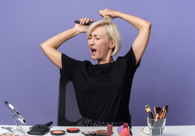 Verärgertes junges schönes mädchen sitzt am tisch mit make-up-werkzeugen, die haare einzeln auf blauer wand kämmen