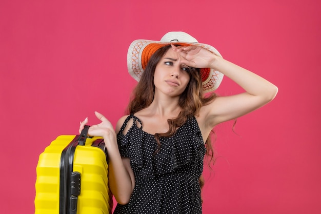 Verärgertes junges schönes reisendes Mädchen im Kleid im Tupfen im Sommerhut, der Koffer hält Kamera mit traurigem Ausdruck auf Gesicht, das über rosa Hintergrund steht