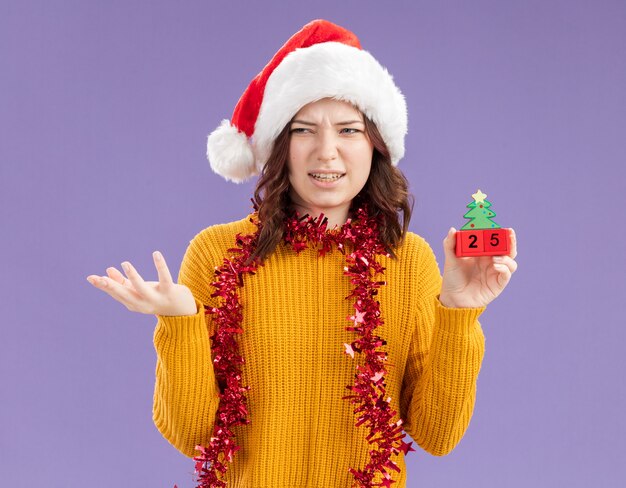 Verärgertes junges slawisches Mädchen mit Weihnachtsmütze und mit Girlande um Hals, der Weihnachtsbaumverzierung hält und Seite lokalisiert auf lila Hintergrund mit Kopienraum betrachtet