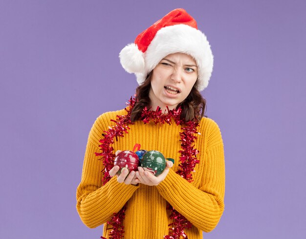 Verärgertes junges slawisches Mädchen mit Weihnachtsmütze und mit Girlande um Hals hält Glaskugelverzierungen, die Seite lokalisiert auf lila Hintergrund mit Kopienraum betrachten