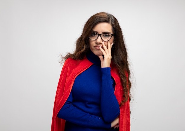 Verärgertes kaukasisches Superheldenmädchen mit rotem Umhang in optischen Gläsern setzt Hand auf Gesicht lokalisiert auf weißer Wand mit Kopienraum