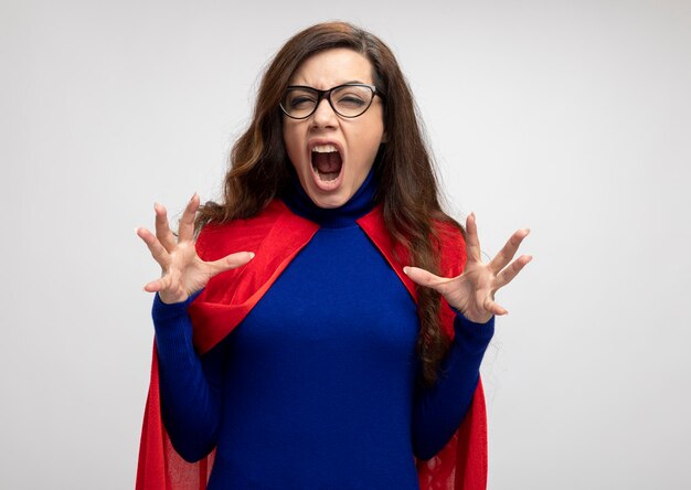 Verärgertes kaukasisches Superheldenmädchen mit rotem Umhang in optischer Brille steht mit erhobenen Händen auf Weiß