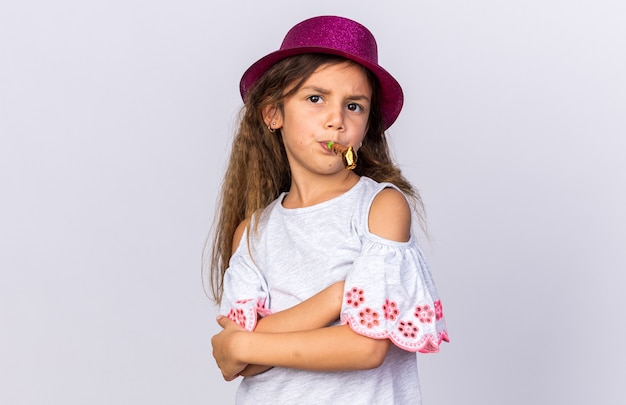 Verärgertes kleines kaukasisches Mädchen mit lila Partyhut, das mit verschränkten Armen steht und Partypfeife bläst, isoliert auf weißer Wand mit Kopierraum