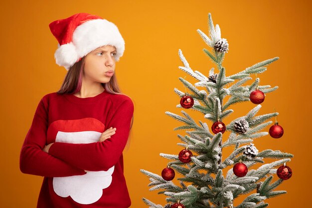 Verärgertes kleines Mädchen in Weihnachtspullover und Weihnachtsmütze, das neben einem Weihnachtsbaum steht und ihn mit stirnrunzelndem Gesicht mit verschränkten Armen auf orangefarbenem Hintergrund betrachtet