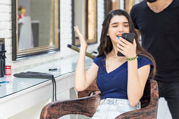 Verärgertes Mädchen, das im Friseursalon sitzt und auf ihr Telefon schaut Hochwertiges Foto