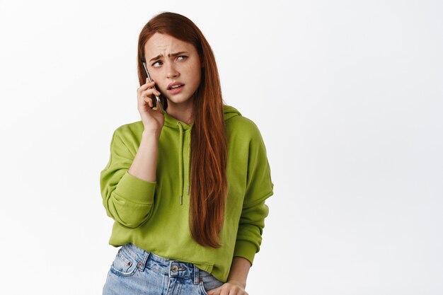 Verärgertes rothaariges Mädchen spricht am Telefon, führt unangenehme verwirrende Gespräche, ruft jemanden auf dem Handy auf Weiß an