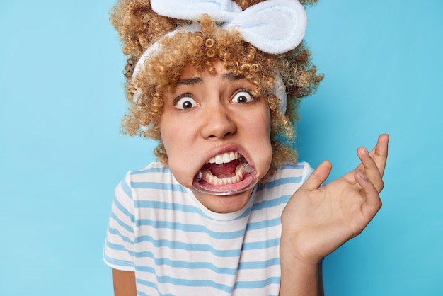 Verblüffte, emotionale, lockige Frau starrt verwanzte Augen an, verwendet einen Mundexpander, hält die Handfläche hoch, trägt ein Stirnband und ein gestreiftes T-Shirt, reagiert auf etwas Peinliches, isoliert vor blauem Hintergrund