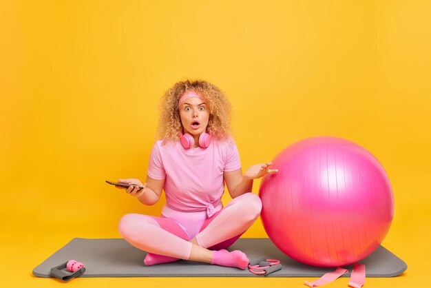 Verblüffte, schockierte Sportlerin in rosa Activewear sitzt in Lotus-Pose auf einer Fitnessmatte und hält ein Mobiltelefon in der Hand. Sie findet heraus, dass schreckliche Nachrichten Fitball-Expander und ein Widerstandsband für das Training im Fitnessstudio verwenden