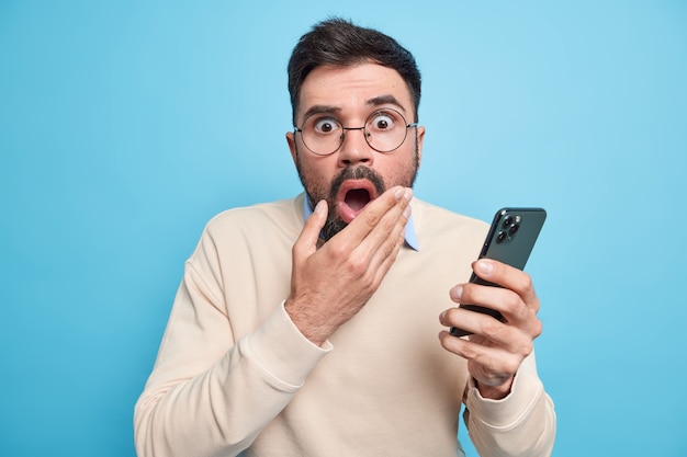 Verblüffter bärtiger erwachsener europäischer Mann hält den Mund offen, starrt erschrocken, hält das Handy und erfährt schockierende Neuigkeiten in einem lässigen Pullover