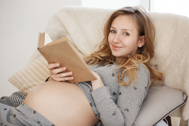 Verbringen Sie wertvolle Zeit damit, die Schwangerschaft bald zu beenden. Porträt der jungen charmanten fröhlichen schwangeren Mutter mit den blonden Haaren, die im Bett liegen und Lieblingsbuch lesen, das sich am Morgen entspannt.