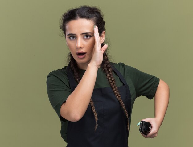 Verdächtige junge Friseurin in Uniform, die Haarschneidemaschinen hält, flüstert isoliert auf olivgrüner Wand