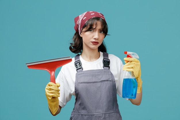 Verdächtige junge Putzfrau mit einheitlichem Bandana und Gummihandschuhen, die Wischer und Reinigungsmittel zeigen, die isoliert auf blauem Hintergrund in die Kamera blicken