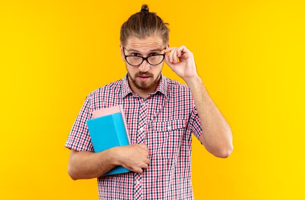 Verdächtige junge Studentin mit Rucksack mit Brille, die Buch isoliert auf oranger Wand hält