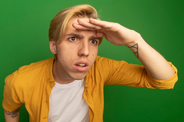 Kostenloses Foto verdächtiger blick auf den jungen blonden kerl der seite, der gelbes t-shirt trägt, das entfernung mit hand lokalisiert auf grün betrachtet