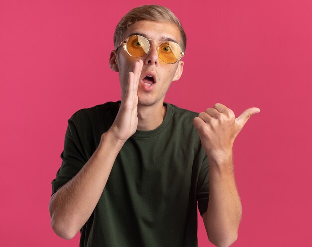 Verdächtiger junger hübscher Kerl, der grünes Hemd und Brille trägt, flüstert und zeigt seitlich isoliert auf rosa Wand mit Kopienraum