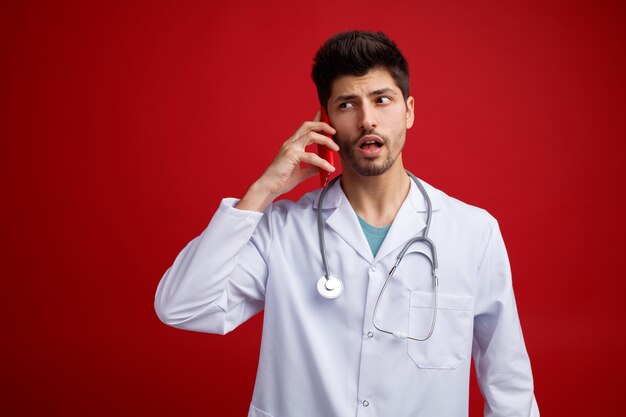 Verdächtiger junger männlicher Arzt mit medizinischer Uniform und Stethoskop um den Hals, der auf die Seite blickt, die am Telefon isoliert auf rotem Hintergrund spricht