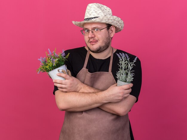 Verdächtiger junger männlicher Gärtner, der Gartenhut trägt, der Blumen in Blumentöpfen hält und kreuzt, die auf rosa Wand lokalisiert werden