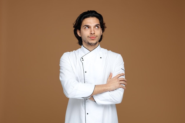 Verdächtiger junger männlicher Koch in Uniform, der auf die Seite schaut und die Hände isoliert auf braunem Hintergrund hält