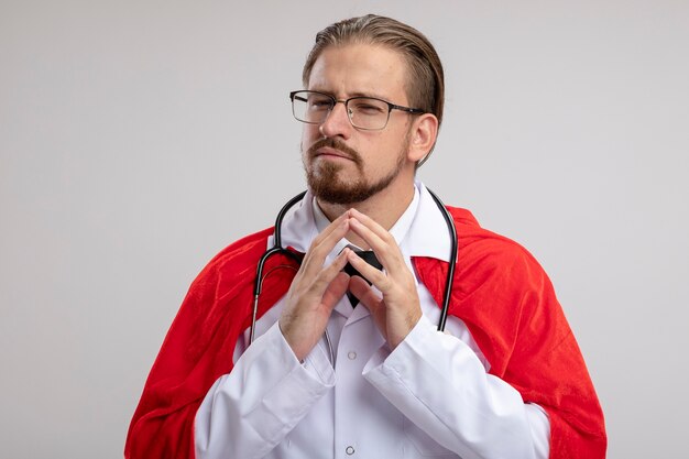 Verdächtiger junger Superheldenmann, der medizinische Robe mit Stethoskop und Brille hält, die Hände zusammen lokalisiert auf weißem Hintergrund halten