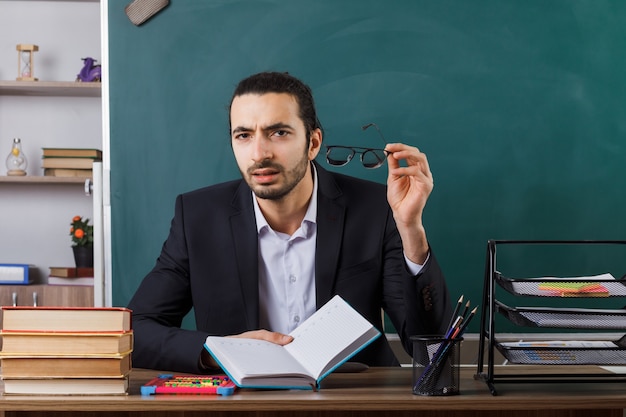 Verdächtiger männlicher Lehrer mit Brille, der ein Buch hält, das am Tisch mit Schulwerkzeugen im Klassenzimmer sitzt