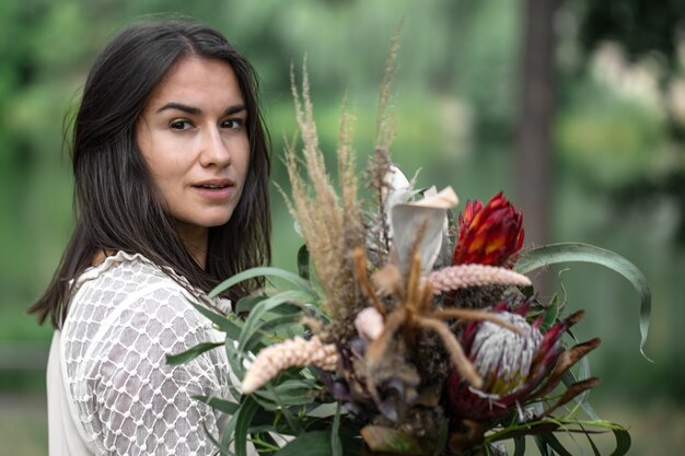 Verführerische junge brünette Frau in einem weißen Kleid mit einem Blumenstrauß im Wald auf unscharfem Hintergrund,