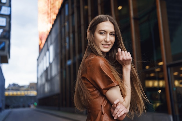 Verführerische und freche junge kaukasische Frau mit langen Haaren, die ein modisches braunes Kleid trägt und eine leere Straße entlang geht.