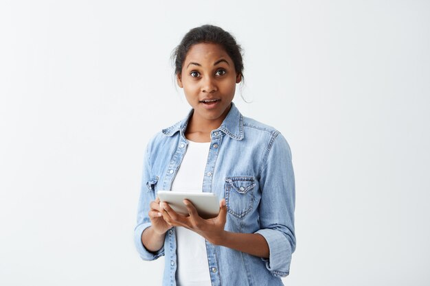 Vergessliche afroamerikanische Frau mit dunklem ansprechendem blauem Hemd, das Tablette in ihren Händen hält und mit erstauntem erstauntem Blick starrt, etwas Ernstes völlig vergessen.