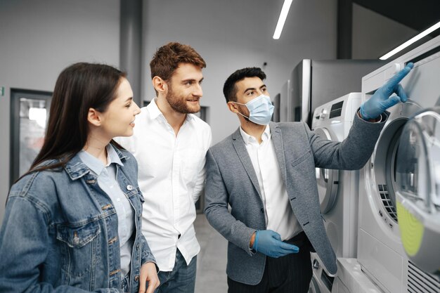 Verkäufer im SB-Warenhaus mit medizinischer Maske zeigt seinen Kunden eine neue Waschmaschine