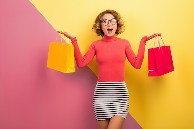 Verlassene attraktive Frau im stilvollen bunten Outfit, das Einkaufstaschen mit überraschtem Gesichtsausdruck, lustiger Emotion, rosa gelbem Hintergrund, Polohals, gestreiftem Minirock, Verkauf, Discout, Shopaholic hält