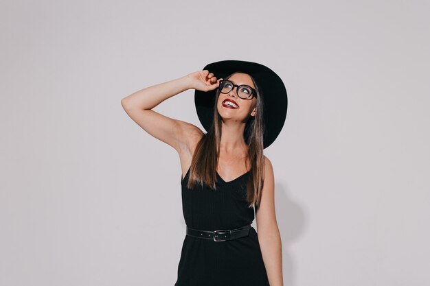 Verlassenes stilvolles glückliches Mädchen, das schwarzes Kleid und Hut hält, die Brille hält und mit Lächeln nach oben schaut