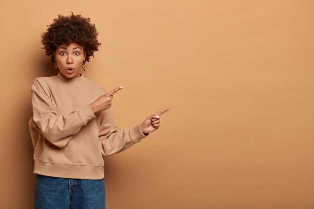 Verlegen erschrockene afroamerikanische Frau zeigt beiseite, schnappt nach Luft, zeigt den Weg und reagiert auf erstaunliche Neuigkeiten, trägt beige Sweatshirt und Jeans, empfiehlt, sich anzumelden und den Online-Shop zu besuchen